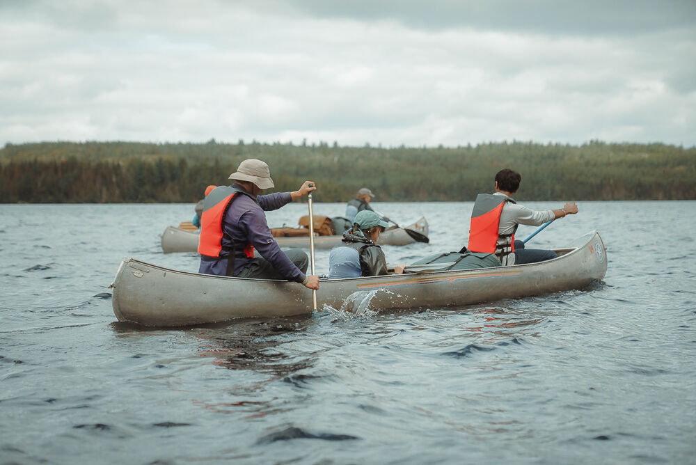 The image shows four people canoeing on a lake. They are all wearing life jackets. The person in the front canoe is wearing a hat. The water is choppy and the sky is cloudy. There are trees in the background.